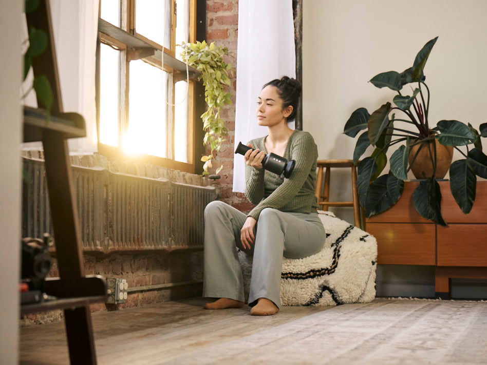 Woman sitting on pouf holding a handheld massager while resting by a sunlit window