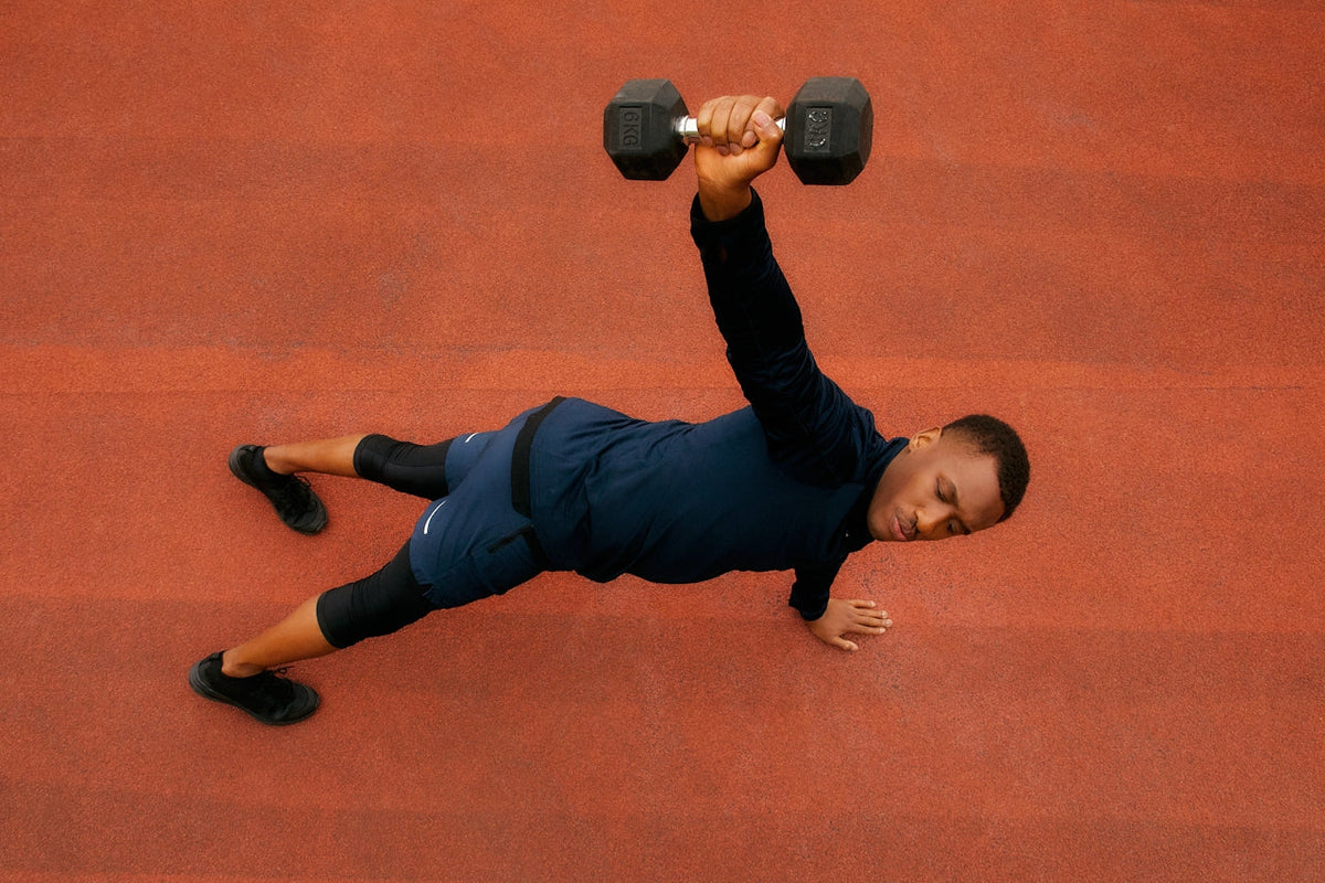 Man performing a side plank on a red track surface, holding a 6 kg dumbbell overhead with one hand, wearing black athletic wear.