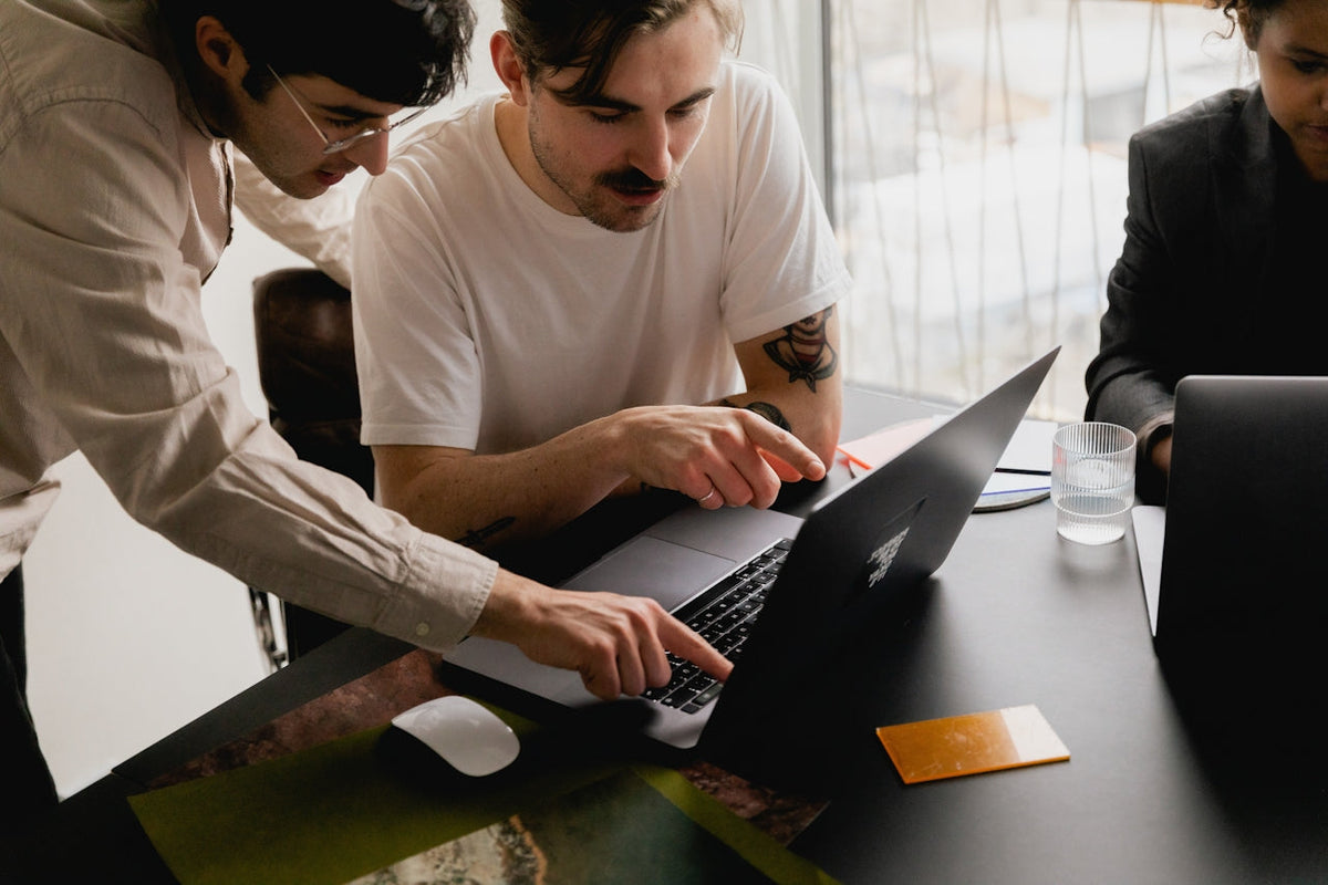 Two men pointing at a laptop screen while collaborating at a table with another person working on a laptop nearby.