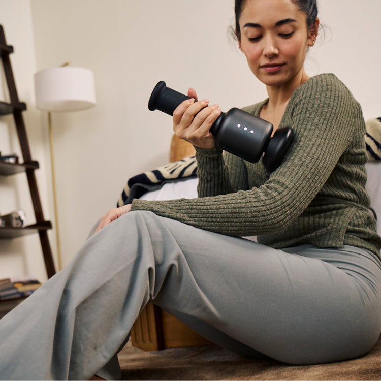 Woman using a black orbital massager on her arm while sitting on the floor in a cozy room
