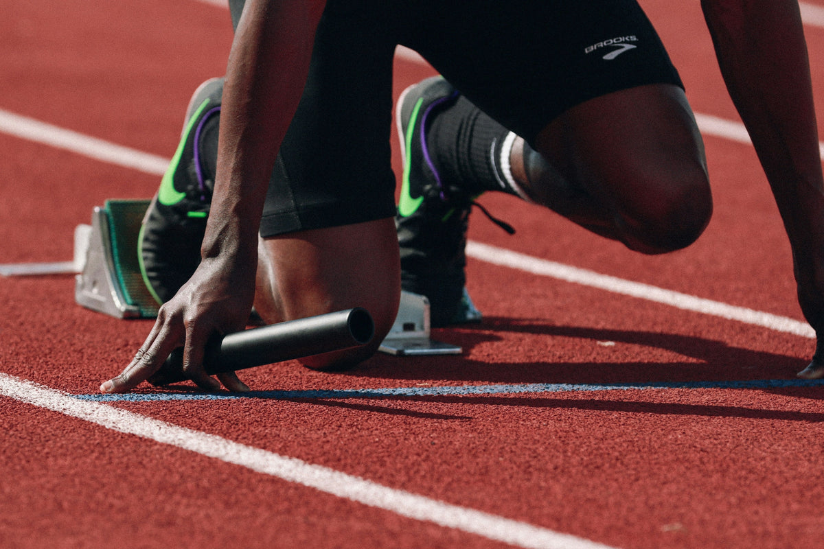 Runner kneeling in starting blocks holding a baton on a red track, wearing shorts labeled 'BROOKS'.