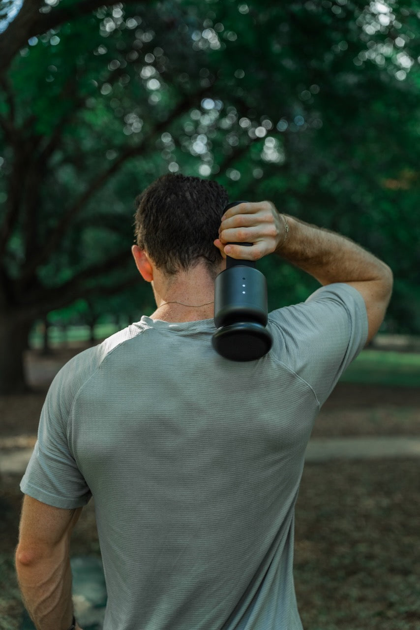 Man seen from behind holding a black percussion massage gun to his upper back/neck in a tree-lined park.