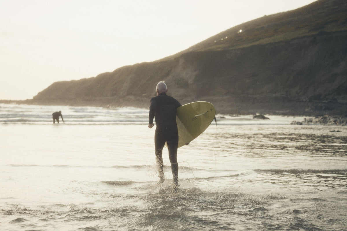 Surfer in a wetsuit carrying a yellow surfboard walking into calm morning ocean with rocky cliffs in the background