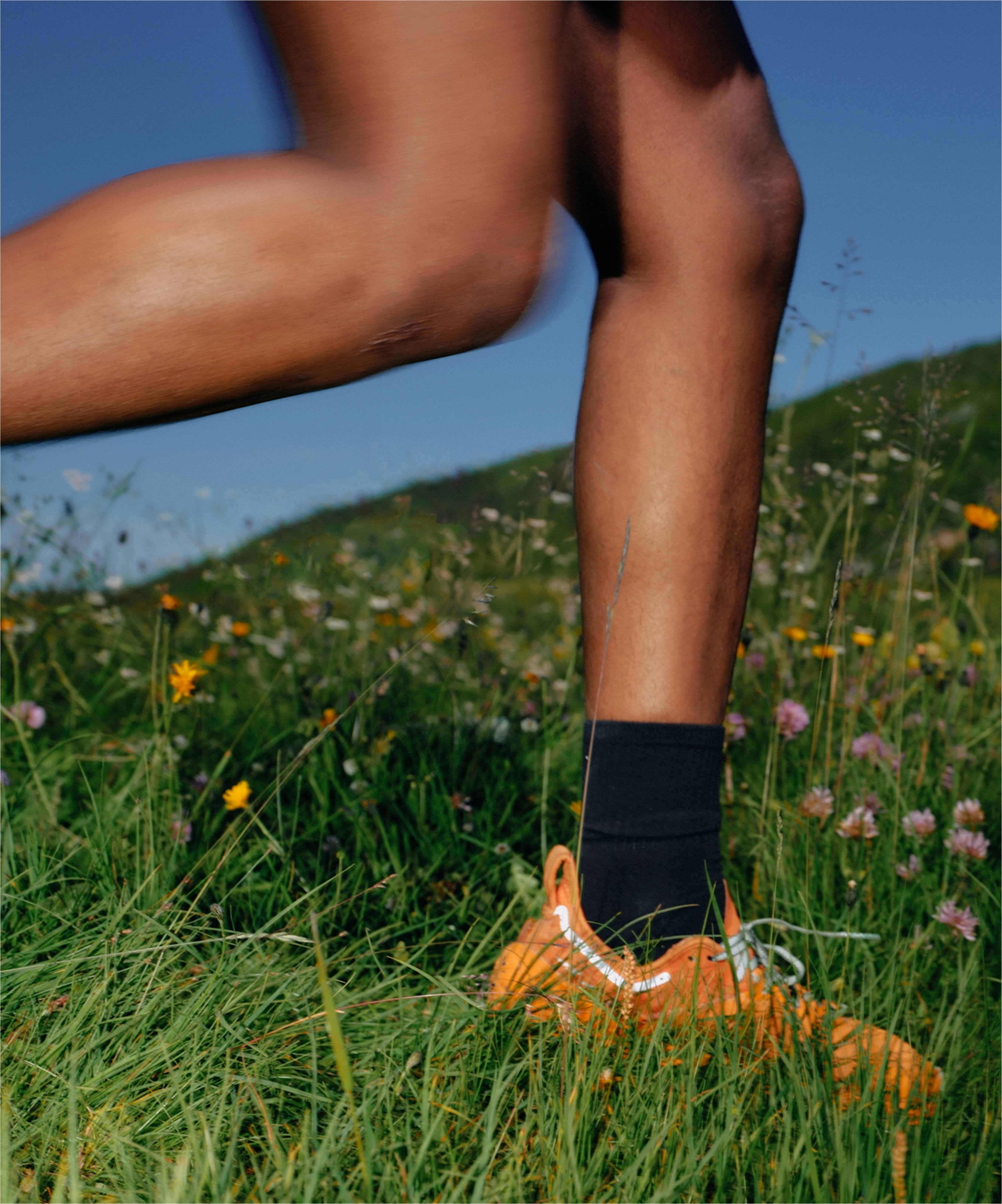 Runner's lower legs and orange trail shoe in mid-stride through grassy meadow with wildflowers and blue sky.
