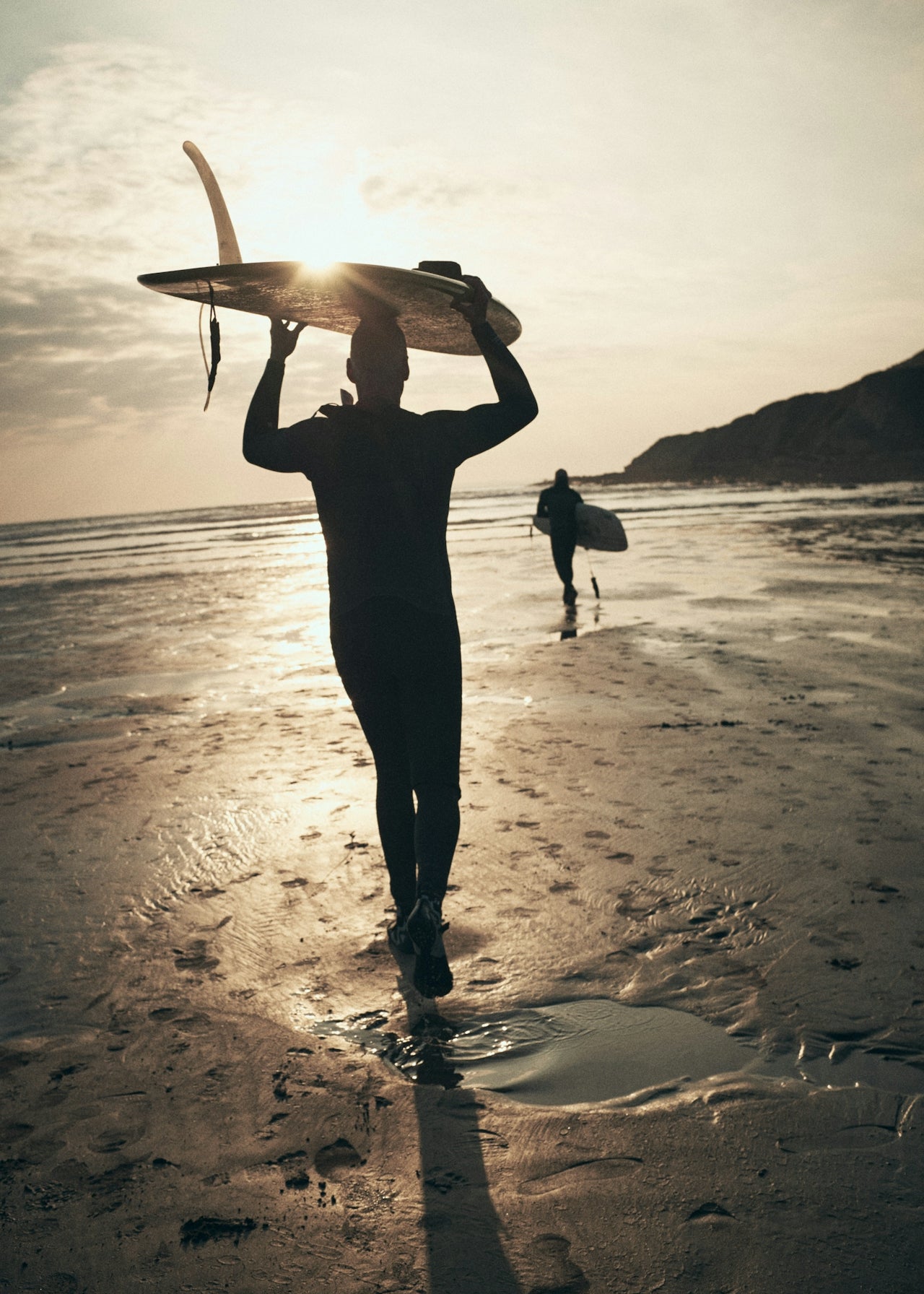 Surfer carrying a surfboard overhead walking across wet sand toward the ocean at sunset