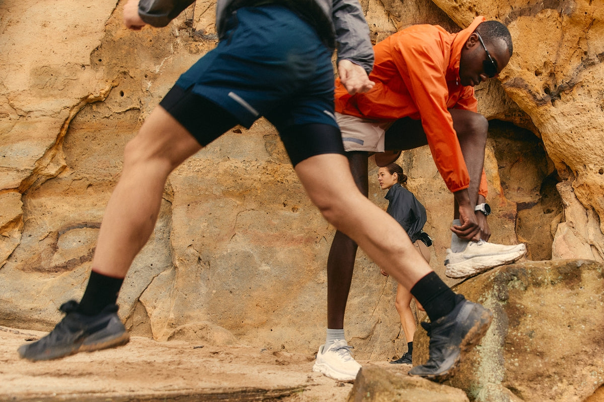 Runners on a rocky trail: foreground runner mid-stride, a man in an orange jacket tying his shoe against sandstone cliffs.