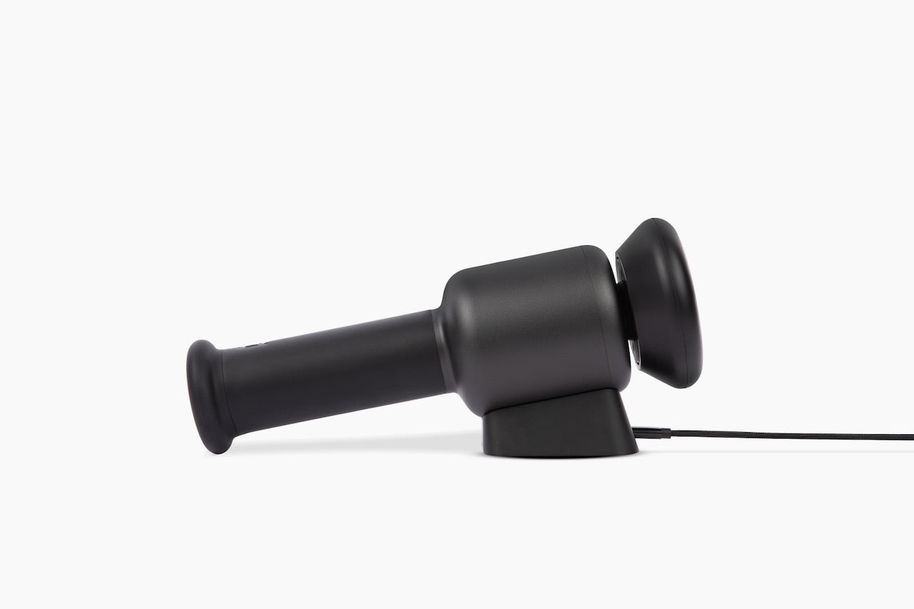 Black handheld percussive massage gun on charging dock, side view with braided charging cable visible.